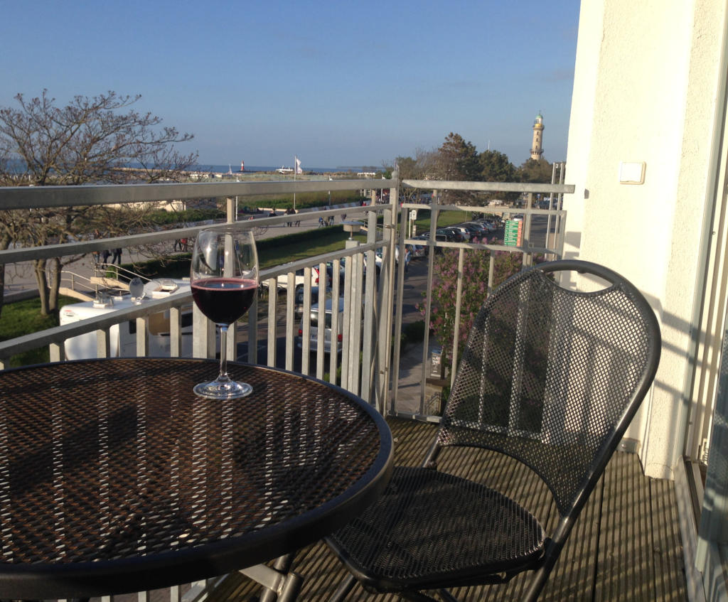 Balkontisch mit einem Glas Rotwein, im Hintergrund die Seepromenade und der weiße Leuchtturm von Warnemünde