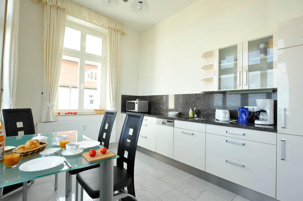 Weiße Einbauküche mit Geschirrspüler, Mikrowelle und Kaffeemaschine, davor ein gedeckter Frühstückstisch am Fenster