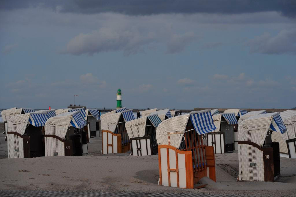 Blau-weiße Strandkörbe im Sand, dahinter das grüne Molenfeuer von Warnemünde