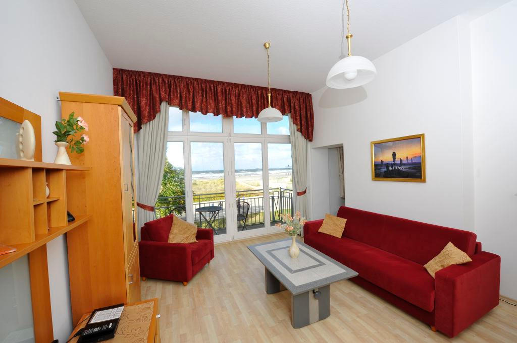 Wohnzimmer mit rotem Sofa, Couchtisch und bodentiefer Balkontür, durch die man Dünen und die Ostsee sieht